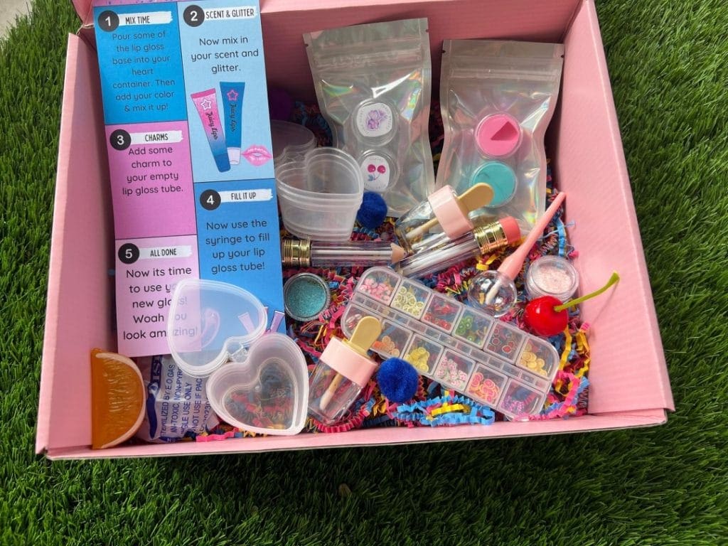 A pink box on grass filled with various DIY lip gloss making supplies including tubes, glitter, charms, and mixing tools for fun delivered.