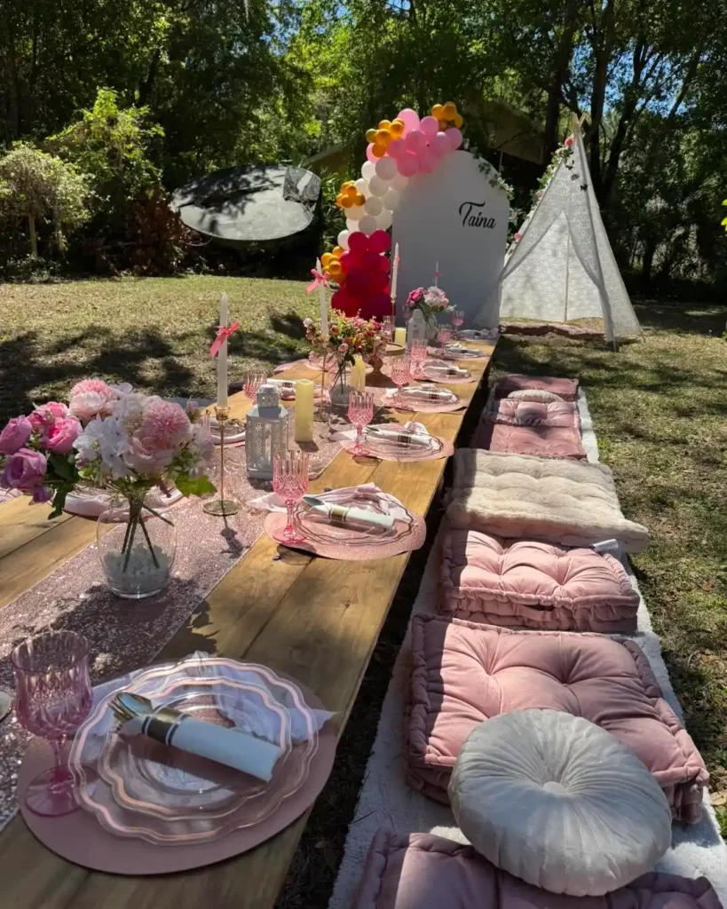 A long outdoor picnic table set with pink and white tableware, flowers, cushions, and balloons stands in a sunny garden with a white teepee and a Shimmer Backdrop Wall Rental in the background.