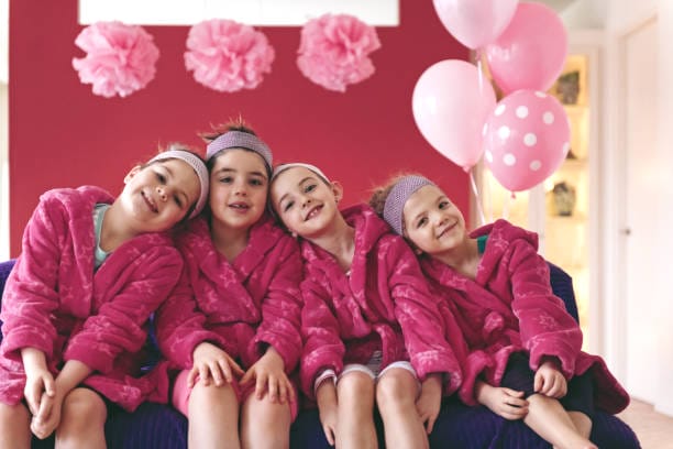 Three young girls in pink robes, with matching headbands, smiling and sitting closely together on a couch, enjoying their DIY Lipgloss Party Box, with pink balloons and decorations in the background.