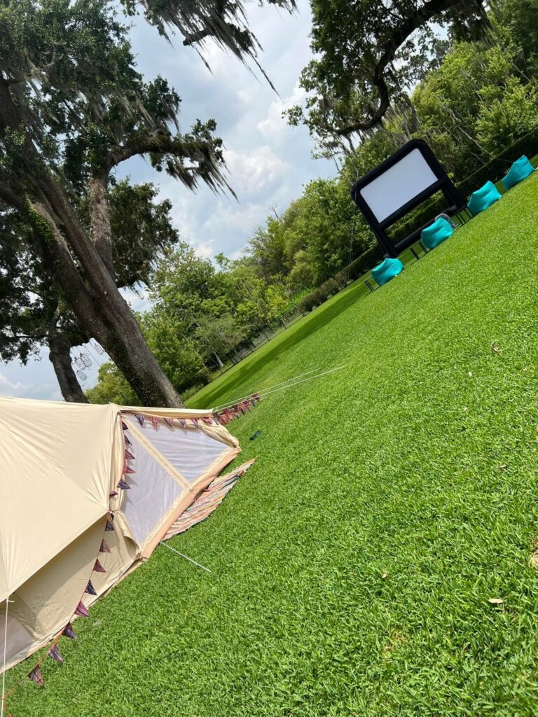 A canvas tent pitched on a grassy lawn in Orlando, with a movie screen and blue chairs set up in the background under hanging moss from trees.