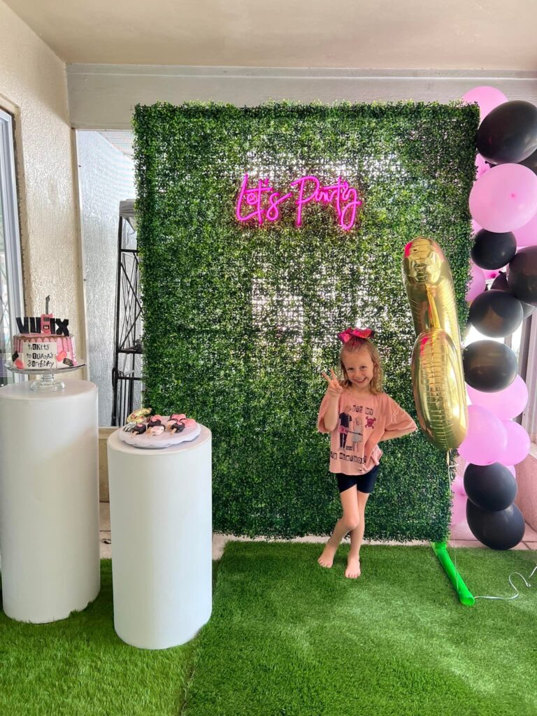 A young girl standing by a "let's party" neon sign on a green hedge wall, decorated with pink and black balloons and an Ultimate Sleepover dessert table to the side.