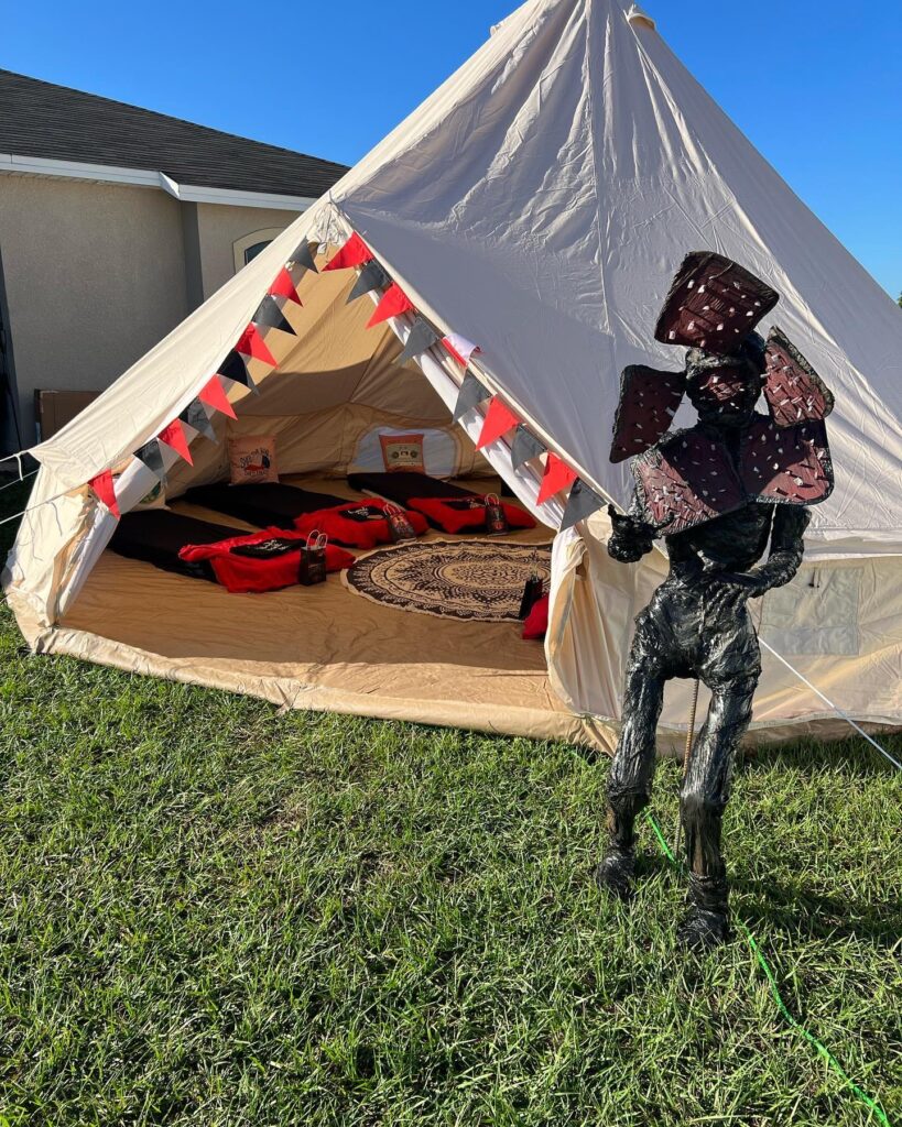 A person in a camouflage suit stands outside a large tent set up on grass, the tent adorned with red and white pennants for the Ultimate Sleepover Experience.