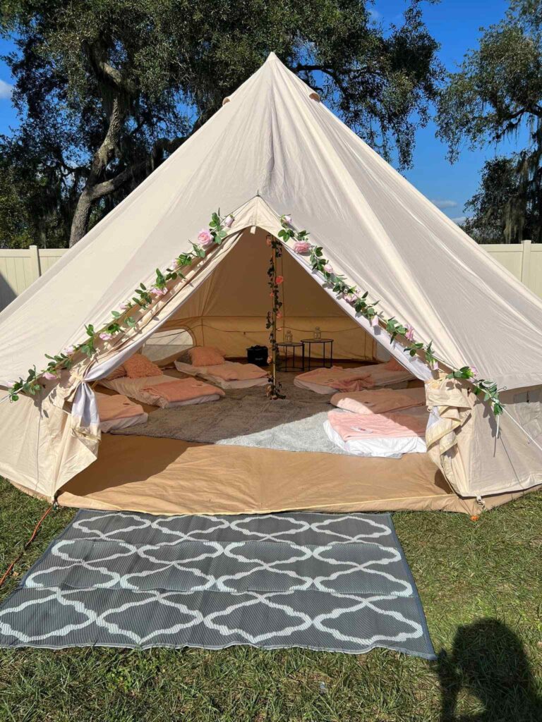 A canvas glamping tent decorated with flowering vines, open to show a cozy interior setup with plush bedding and cushions, set against a backdrop of trees in Central Florida and a clear blue sky.
