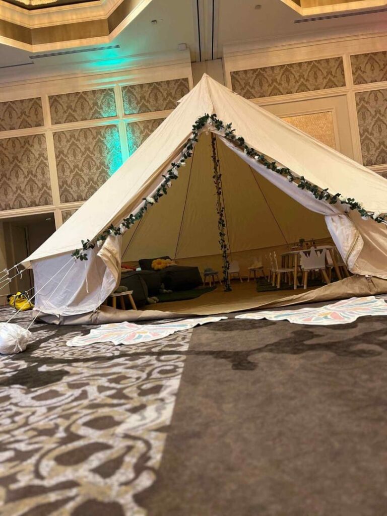 A large indoor glamping-style tent decorated with greenery, set up in a room with elegant wallpaper and patterned carpet for a birthday celebration.