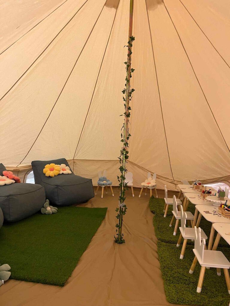 Interior of a large tent with artificial grass flooring, grey sofas, and a dining area with white chairs and a table, decorated with a vine climbing a central pole for an outdoor glamping experience