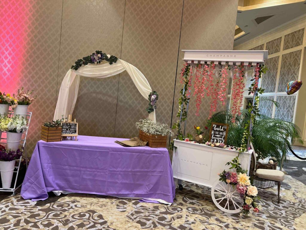 Wedding venue setup with a purple draped table, white floral cart, and a champagne stand under a floral arch for an ultimate sleepover.