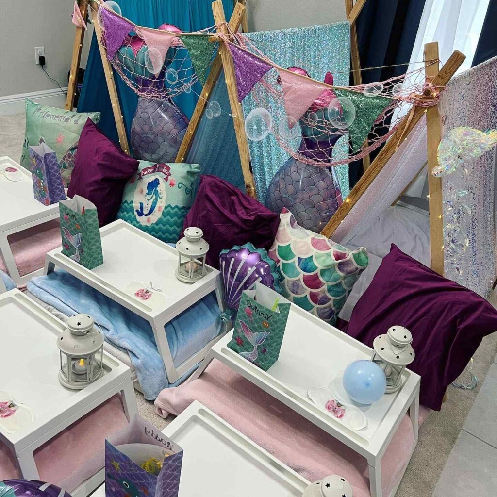 Indoor children's party setup with mermaid theme, featuring decorated teepees for the Ultimate Sleepover experience, cushions, and small tables with lanterns and themed accessories.