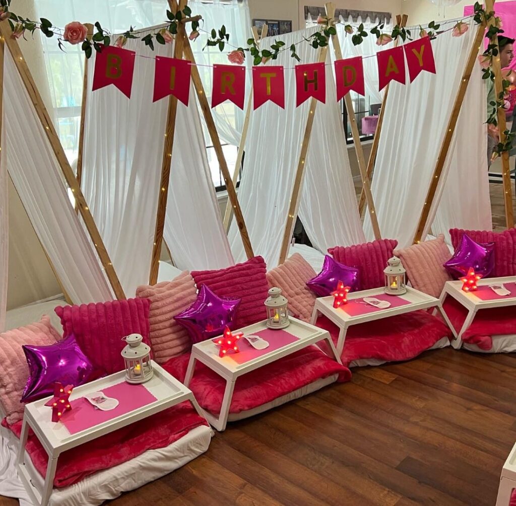 Indoor Slumber Party setup with four small tents draped in white, decorated with pink cushions, star-shaped pillows, and small tables, under a 'birthday' banner.