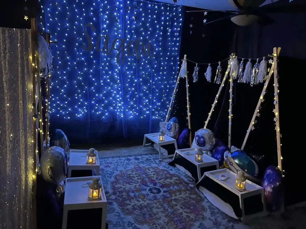 A dimly lit room decorated with a "dream" sign in blue lights, featuring small teepees and celestial-themed cushions on the floor, evoking a magical night.