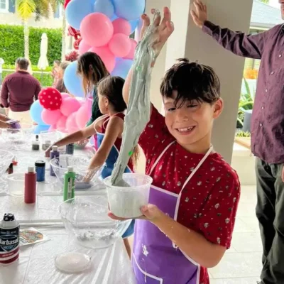 A young boy in a purple apron smiles while lifting a stretchy slime mixture at a crafts table during an outdoor kids party in Central Florida, with balloons in the background.