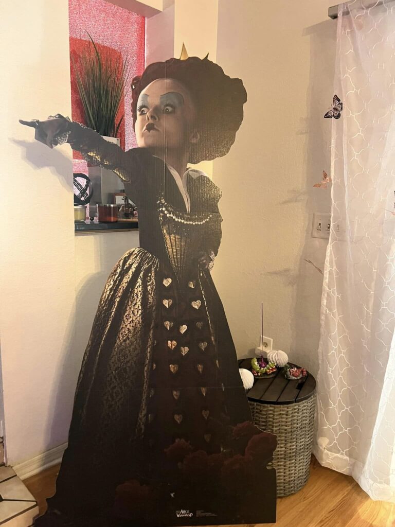 Cardboard cutout of a woman dressed in an elaborate Victorian gown and hat, pointing outward, standing in a Wonderland Wonders party interior.