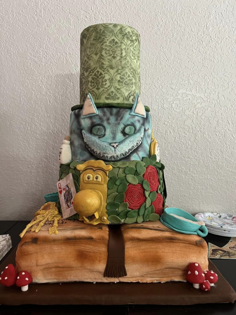 A multi-tiered cake designed for an Alice in Wonderland Party, looking like the Cheshire Cat with a large hat, a pocket watch, and detailed decorations on a wooden-like base.