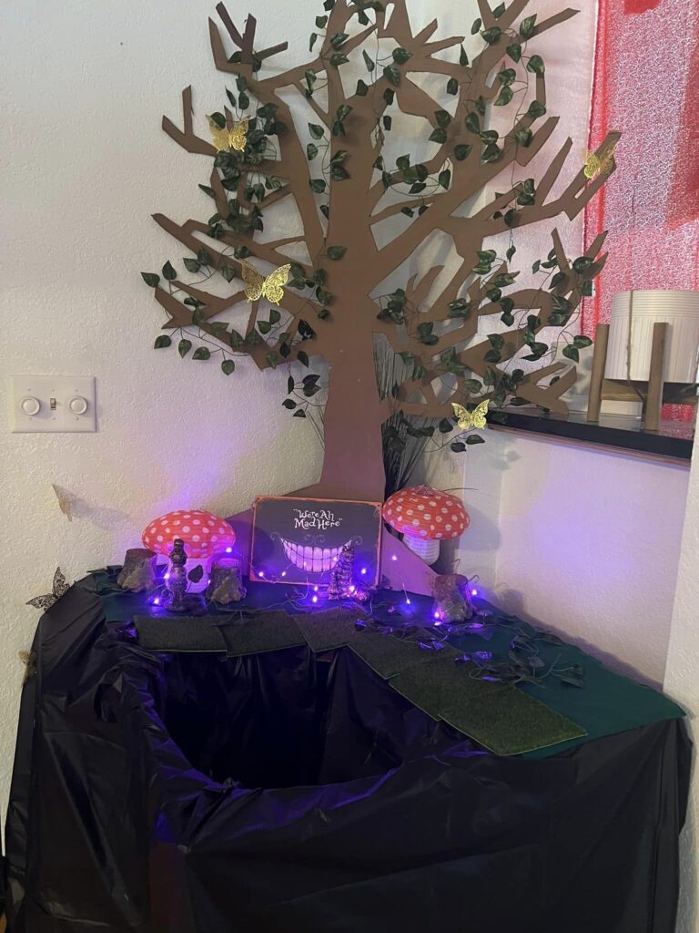 A decorated display straight out of an Alice in Wonderland extravaganza features a handmade tree adorned with paper butterflies, artificial grass, and whimsical mushroom decorations, all illuminated by enchanting purple lights. A "Magic Meadow" sign graces the backdrop.