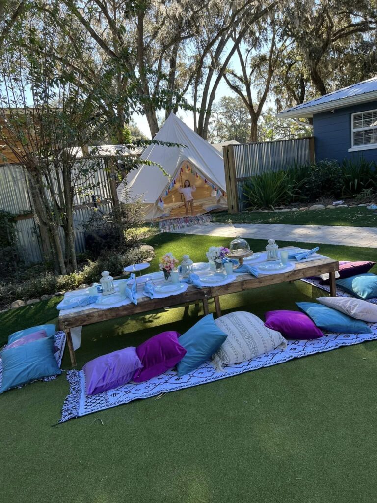 Outdoor children's party setup with a decorated low table, colorful cushions on a rug, and a white tent in a garden for the Ultimate Slumber Party.