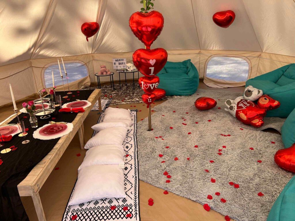 A romantic tent setup for the Ultimate Night, with a decorated table, red heart balloons, plush toys, and rose petals scattered on the floor.