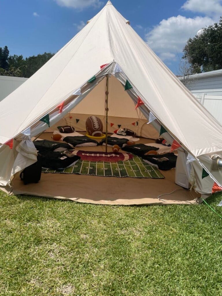 A large white tent set up outdoors with its flaps open, revealing an ultimate slumber party-themed interior with a football rug, bean bags, and festive bunting.
