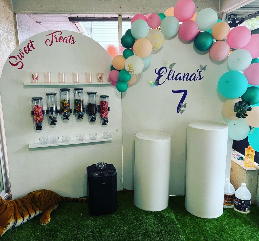 A birthday party setup featuring a candy wall labeled "sweet treats," a balloon arch in pastel colors, and decorations for Eliana's 7th birthday—now upgraded to the 2024 Ultimate