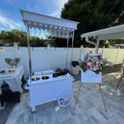 Outdoor catering setup with a white food cart, floral decoration, and an easel sign stating "Elevate Your Party" under a clear blue sky.