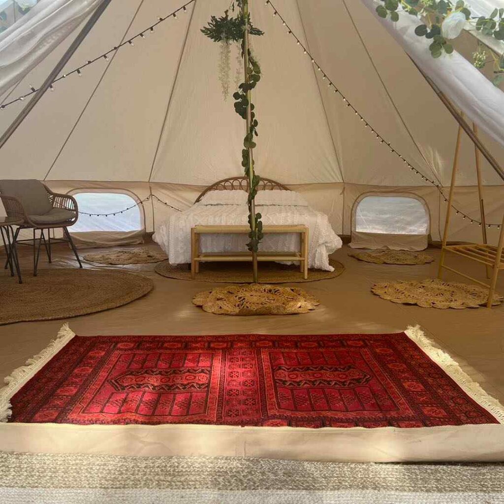 Interior of a Slumberr glamping tent with a plush bed, rich red rug, decorative plants, and open windows, creating a cozy, luxurious camping atmosphere.