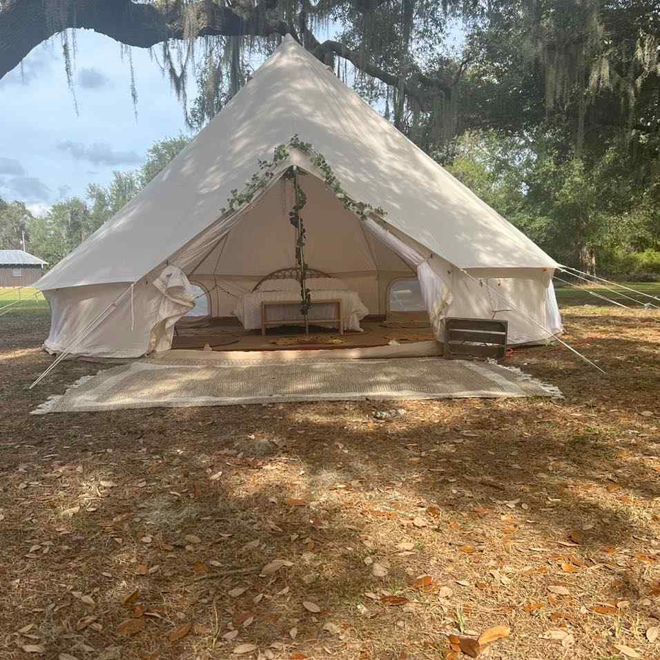 A canvas glamping tent decorated with a floral wreath, partially opened to reveal beds inside, set up in a wooded area with scattered leaves on the ground for an ultimate night.