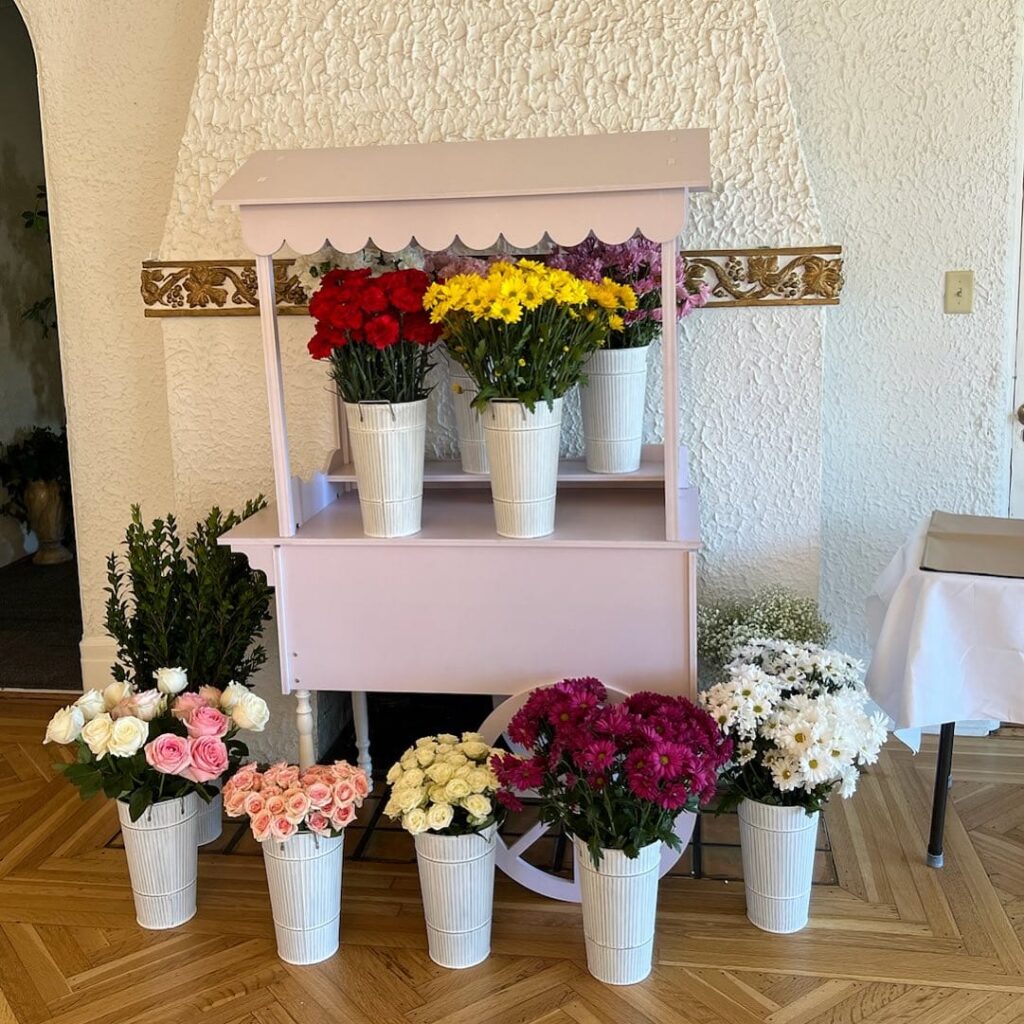 A small, decorative flower cart displaying buckets of colorful flowers including red, yellow, pink, and white blooms, perfect to upgrade your party décor.