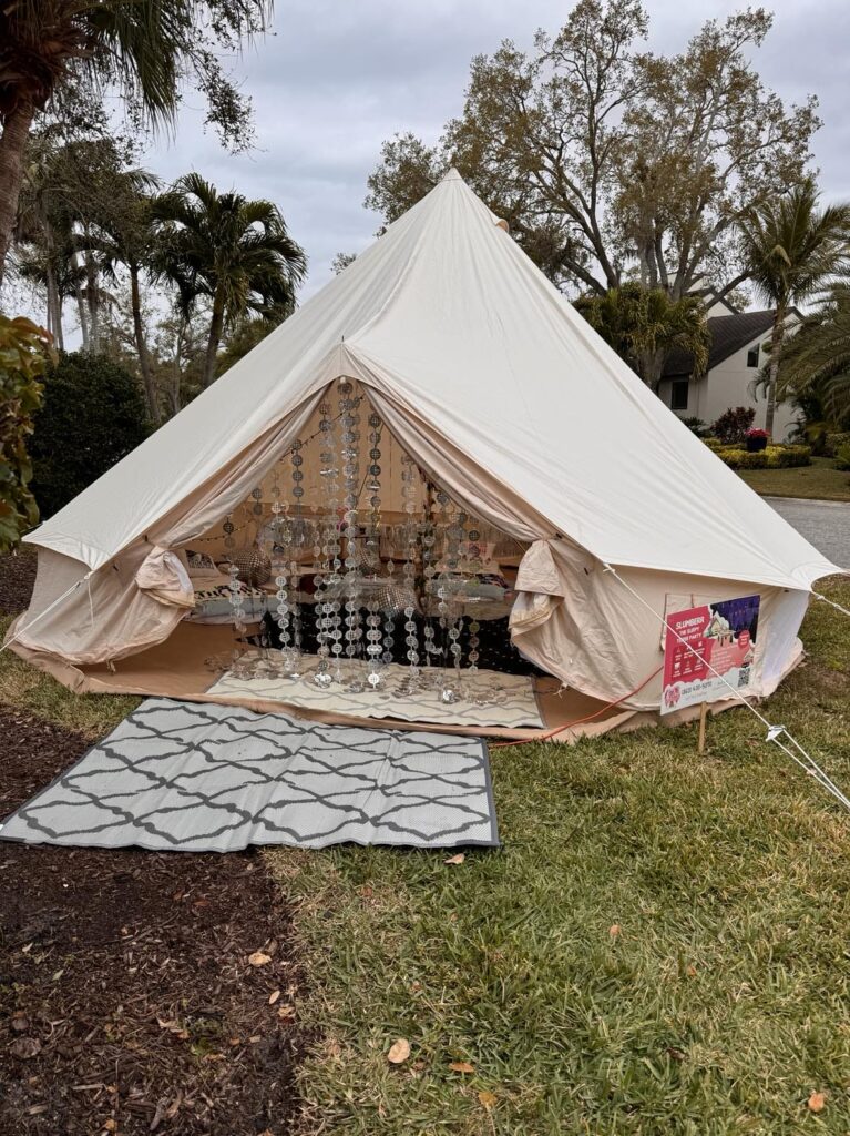 A bell tent set up in a garden with an open entrance, decorated with a chandelier and rugs, surrounded by palm trees for the 2024 Ultimate Sleepover.