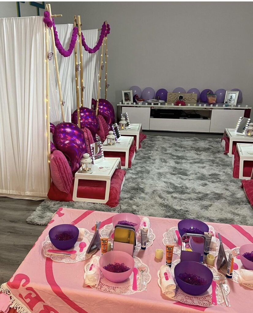 A colorful spa-themed party setup for children with pink and purple chairs, tables, and balloons in a room with white curtains and a gray rug for the 2024 Ultimate Sleepover.