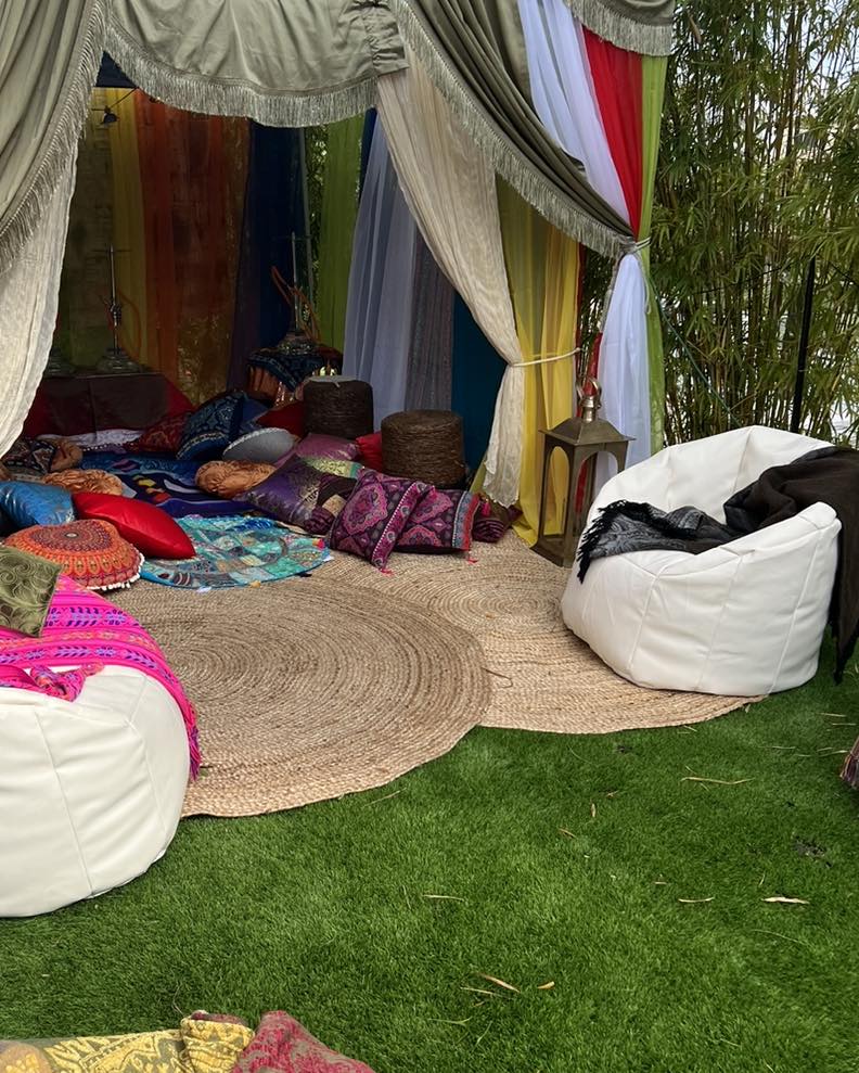 A cozy outdoor seating area with colorful cushions, drapes, and a round rug, set up under a tent-like structure surrounded by lush greenery, perfect for an ultimate night adult party.