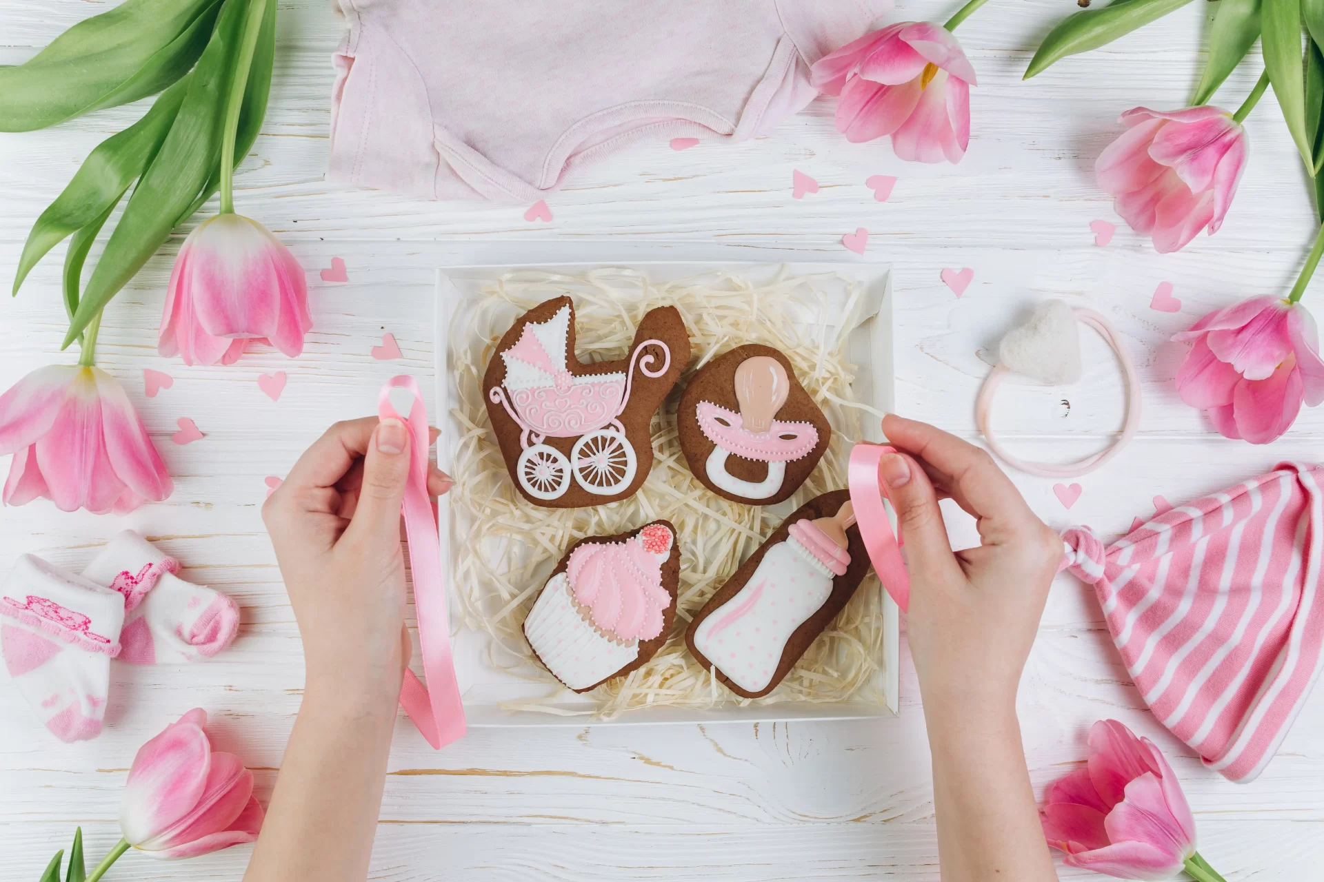 Hands arranging a gift box with baby-themed cookies and small clothes, surrounded by pink tulips on a white wooden table for a onesie painting party.