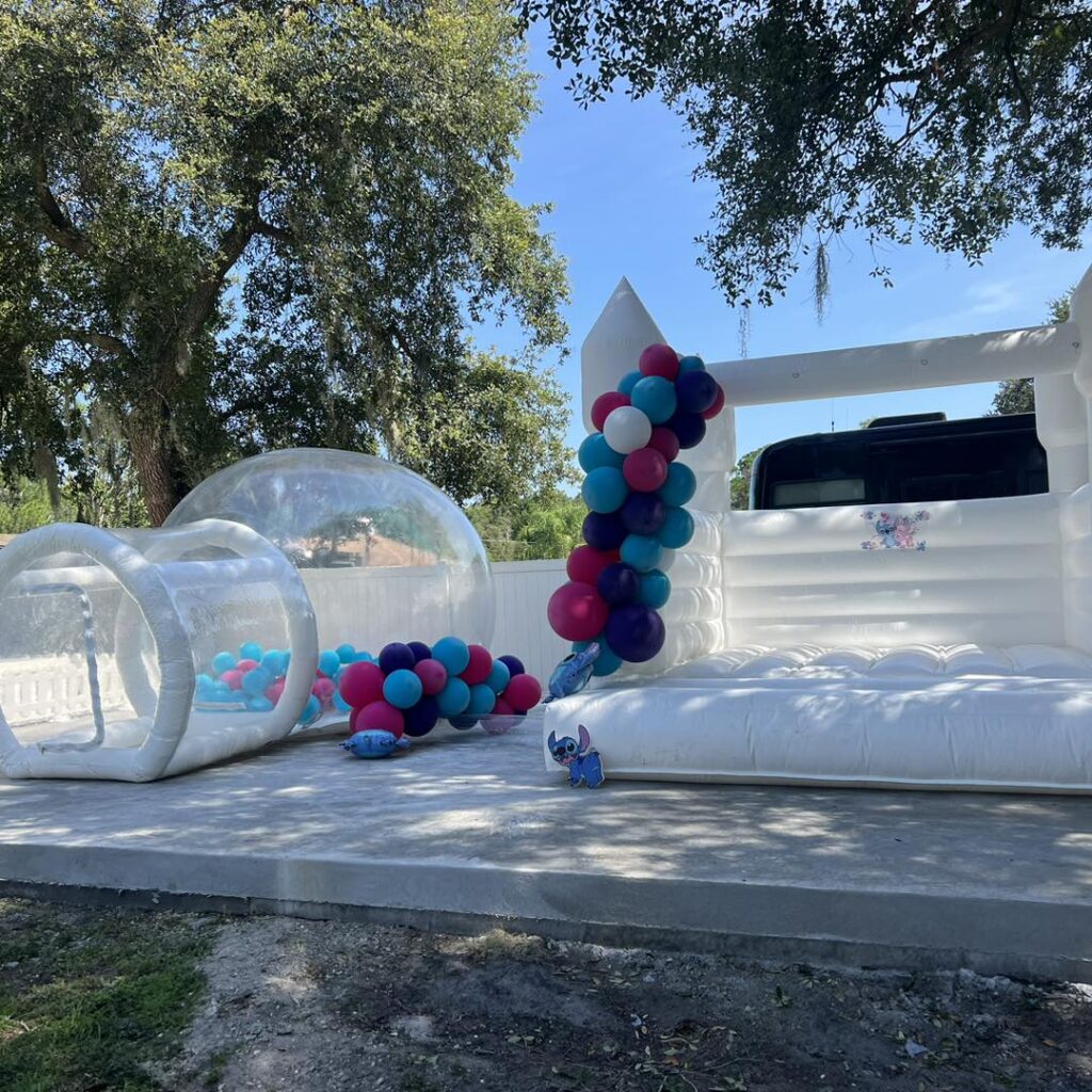Discover the Ultimate Baby Shower Rentals setup featuring a captivating white inflatable bounce house paired with mesmerizing transparent spheres. This charming outdoor scene is adorned with an enchanting blend of blue, pink, purple, and white balloons that add a splash of color and fun. Set against a picturesque backdrop of lush green trees under a vibrant, sunny sky, this delightful arrangement promises to elevate any baby shower experience. Perfect for creating lasting memories and capturing Instagram-worthy moments, this curated rental ensemble is the ideal centerpiece for your celebration.