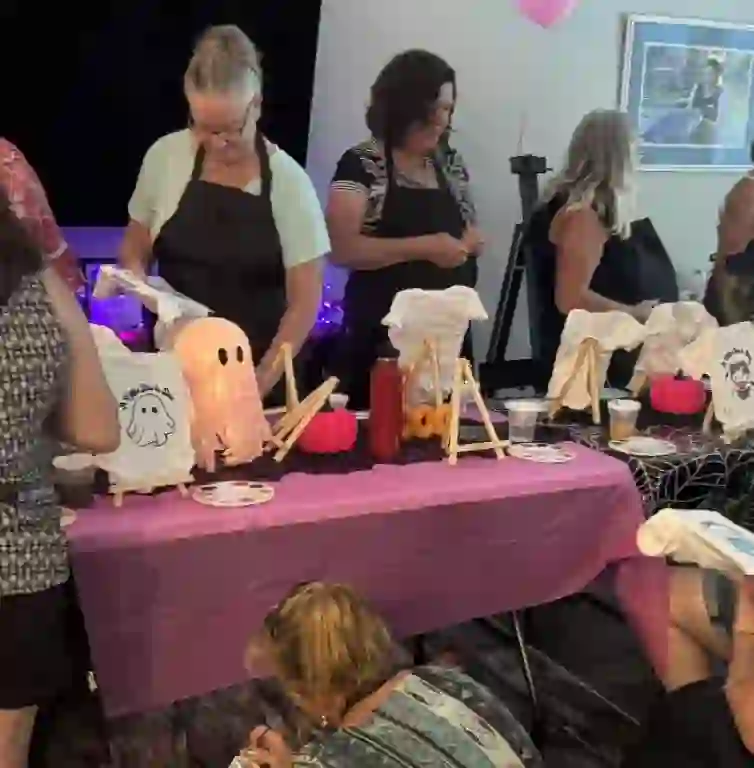 A lively group of participants is gathered around a whimsically decorated table, immersed in a Halloween-themed painting extravaganza that evokes the charm of an ultimate baby shower rental setup. The scene is set with a soft pink cloth covering the table, which is adorned with intricately designed pumpkins, inviting creative spirits to unleash their artistic flair. Guests enthusiastically paint ghostly designs on fabric, adding to the festive ambiance. The enchanting atmosphere is further elevated by captivating Halloween decorations, creating a perfect blend of creativity and celebration. Keywords: Halloween painting activity, baby shower rental setup, decorated pumpkins, ghost design fabric painting, festive decor atmosphere.
