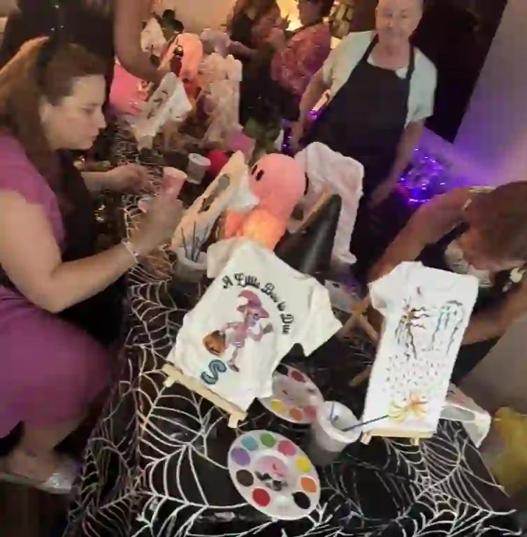 Engage in Creativity: A Lively Craft Table Scene with Spider Web Tablecloths and Vibrant T-Shirt PaintingCapture the essence of creativity at a bustling craft table, adorned with festive spider web tablecloths perfect for Halloween or themed events. In the foreground, a woman skillfully wields a paintbrush, bringing designs to life on an array of colorful T-shirts. Her focus is matched by several others who gather around, each contributing to the artistic atmosphere. Paint palettes and cups speckle the scene, enhancing the vibrant palette of this interactive experience. Decorative lighting in the background casts a warm glow over this lively setting, ideal for those seeking unique activities from Ultimate Baby Shower Rentals. This picture-perfect moment reflects not only collaborative spirit but also showcases innovative T-shirt designs that add flair to any gathering.