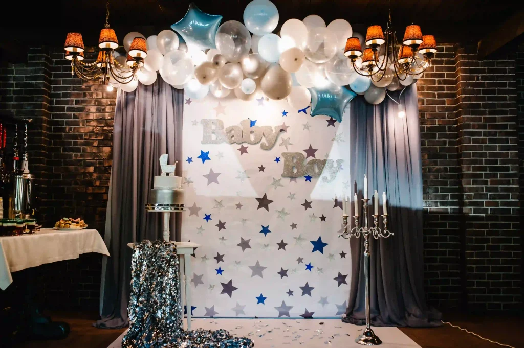 Experience the ultimate baby shower celebration with this captivating setup designed to welcome a baby boy. The elegant display features a charming "Baby Boy" banner set against a pristine white backdrop, beautifully adorned with shimmering silver and vibrant blue stars. Above the banner, an eye-catching arch of balloons in shades of white, silver, and blue creates a stunning focal point that adds an element of festive whimsy to the room. At the heart of this enchanting arrangement is a magnificent three-tiered cake exquisitely decorated with metallic accents, sitting gracefully on a table which also showcases an ornate silver candlestick. Overhead, warm chandeliers cast a cozy glow over the scene, adding to the inviting ambiance perfect for celebrating this joyous occasion. Perfectly combining creativity and elegance, this unforgettable baby shower setup promises to make your gathering truly special while setting trends in event styling and decor ideas for baby showers welcoming boys.