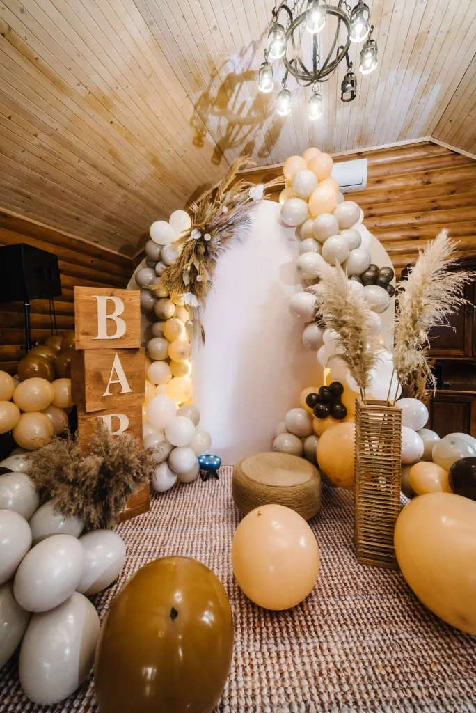 Immerse yourself in the enchanting atmosphere of an Ultimate Baby Shower Rentals setup, where rustic charm meets modern elegance. This inviting wooden room, adorned with a sophisticated balloon arch in soft neutral tones, warmly embraces a pristine white backdrop. Majestic large wooden blocks proudly spell out "BABY," creating an eye-catching centerpiece that draws focus. Lush pampas grass gracefully rises from tall vases, adding texture and an earthy touch to the décor. A charming woven stool invites you to pause and absorb the scene, while artfully scattered balloons infuse playfulness into this serene setting. Perfect for capturing unforgettable memories at any baby shower event!