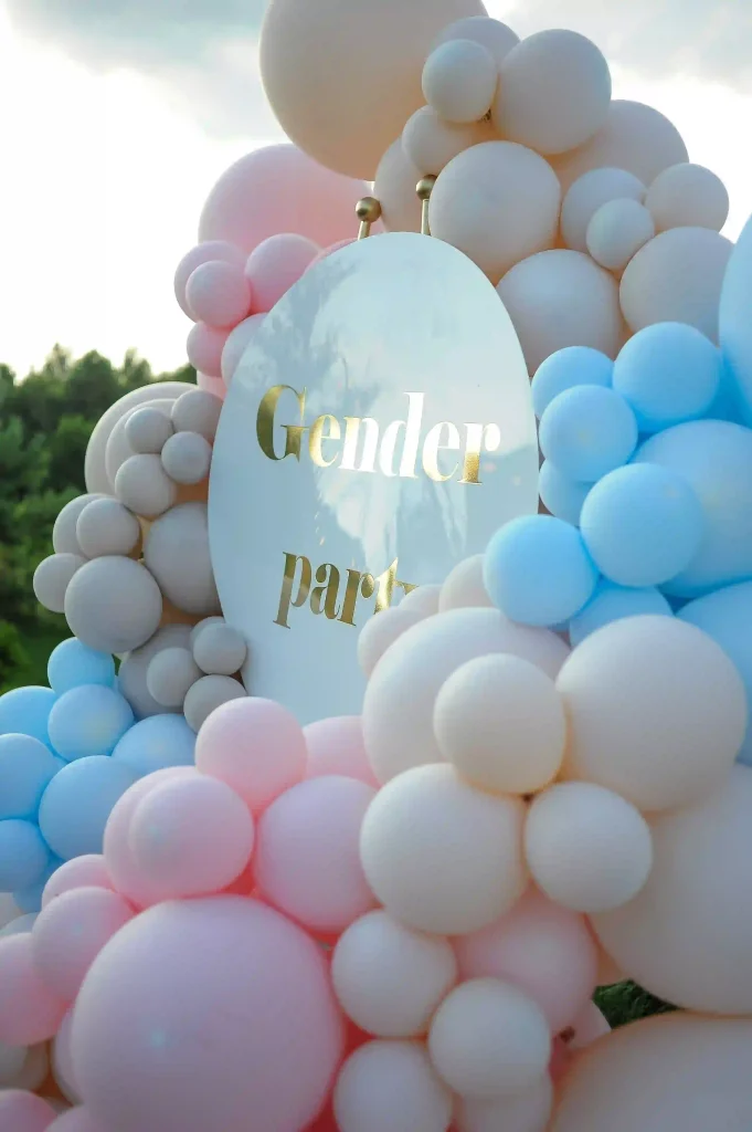 Experience the ultimate baby shower rentals with a stunning gender reveal party setup that captivates and delights! Imagine clusters of vibrant pink, calming blue, and elegant cream balloons artfully arranged around an exquisite oval sign adorned with shimmering reflective gold letters spelling out "Gender Party." These enchanting balloon arches gracefully frame a picturesque backdrop of lush green trees under a partly cloudy sky. Perfect for expecting parents planning unforgettable gender reveal celebrations, this setup combines aesthetic charm with nature’s serene beauty. Discover the ideal blend of creativity and sophistication for your next memorable event.