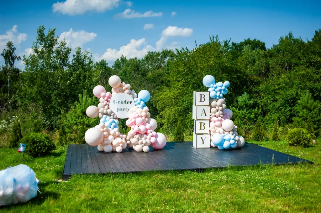 Discover an enchanting gender reveal celebration meticulously arranged by Ultimate Baby Shower Rentals. This captivating outdoor setup features a charming display of pastel pink, blue, and white balloons adorning a rustic wooden platform. The scene is anchored by impressive oversized letter blocks that spell "BABY," creating a perfect photo-op moment. Adding to the excitement is an elegantly crafted sign that reads "Gender Party." Set against a picturesque backdrop of lush green landscape under a vibrant blue sky dotted with fluffy clouds, this dreamy setting offers the ideal environment for celebrating life's joyous milestones. Perfect for those searching for unforgettable gender reveal inspiration!