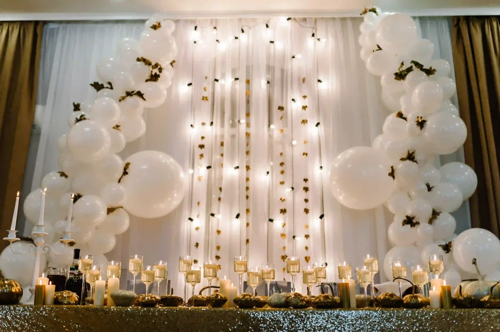Elevate your baby shower experience with the ultimate event rentals that feature a stunning decorated backdrop. This captivating display boasts oversized white balloons, flowing translucent curtains embellished with twinkling fairy lights, and shimmering gold stars. Set against this enchanting scene is a luxurious table adorned in a glittering gold tablecloth, showcasing an array of beautifully lit candles nestled in elegant glass holders. The sophisticated arrangement creates an unforgettable and elegant atmosphere perfect for any memorable celebration. Ideal for those looking to enhance their baby shower decor with elegance and charm.