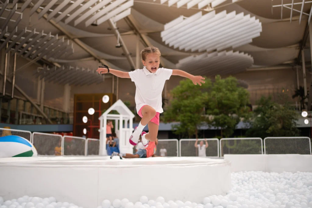 A young girl in a white shirt and pink shorts jumps energetically into a ball pit inside an indoor play area designed for a Sensory-Friendly Sleepover, with people and a playhouse in the background.