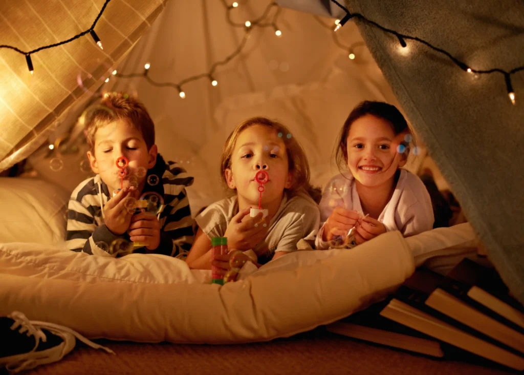 Three children lie inside a blanket fort decorated with string lights, blowing bubbles and smiling, surrounded by books and pillows for a cozy Sensory-Friendly Sleepover.