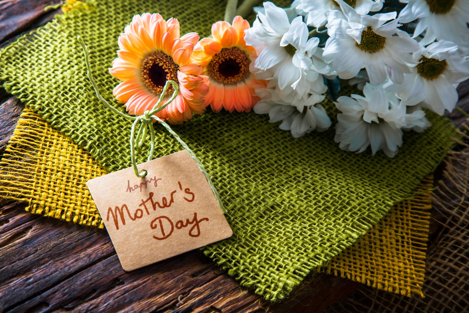A small card reading "Happy Mother's Day" is tied with string next to orange and white flowers, creating a festive Mother's Day display on textured green and yellow fabric atop a wooden surface.