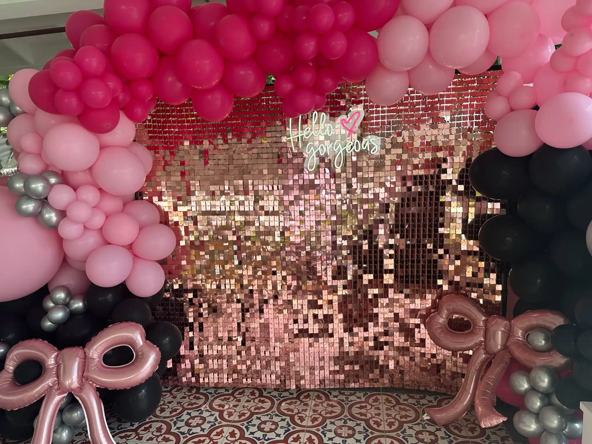 Balloon arch with pink, black, and silver balloons framing a pink sequin backdrop with a "Hello Gorgeous" sign and two large pink balloon bows on a patterned tile floor—perfect for glamping parties or tent-themed kids activities.