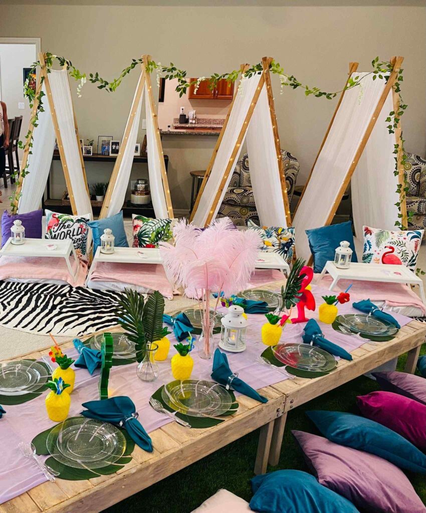 A low wooden table is set with clear plates, teal napkins, and pineapple cups from a South Florida party shop, surrounded by colorful pillows. Small tents with greenery garlands are set up in the background.