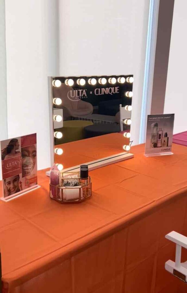A vanity mirror with lights, Clinique and Ulta branding, and assorted makeup products sits on an orange tablecloth—perfect for any South Florida party shop. Promotional displays are placed on either side of the mirror.