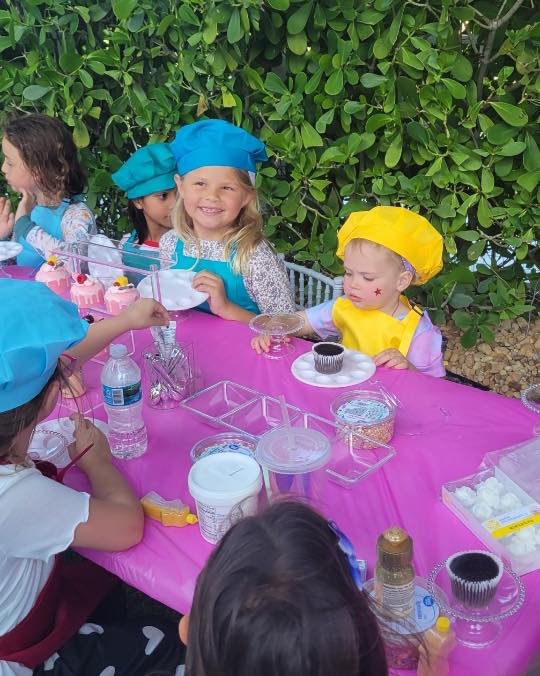 Children wearing colorful chef hats sit around a pink table outdoors, decorating cupcakes with various toppings and supplies at a festive party shop in South Florida.