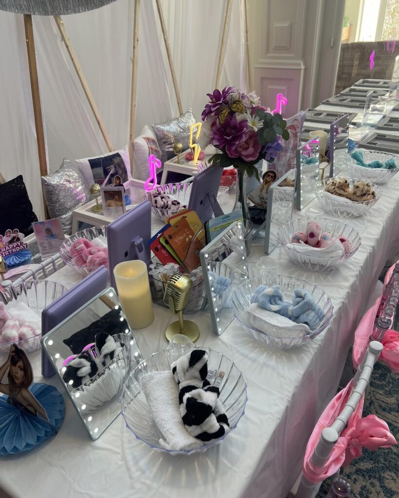 A long table set for a themed kids activity party, with mirrors, bowls of rolled towels, headbands, faux candles, flowers, and decorative items neatly arranged at each seat.