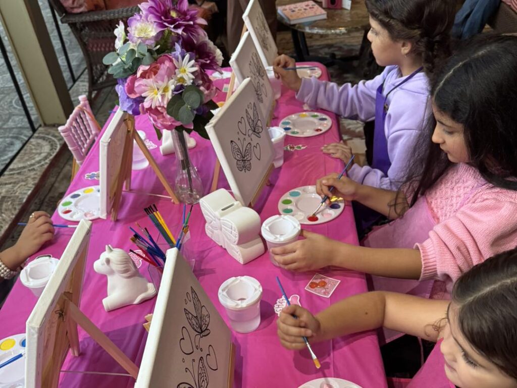 Children sit at a pink-covered table painting butterfly designs on canvases, with paint palettes, brushes, and a vase of flowers in the center—a charming scene perfect for any party planner organizing a creative glamping or teepee-themed celebration.