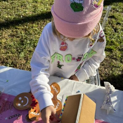 A child wearing a pink beanie and white sweatshirt enjoys a fun kids activity, decorating gingerbread cookies and a gingerbread house at an outdoor table.