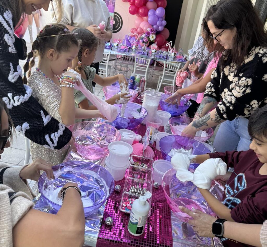 Children and adults gather around a table making tie-dye crafts with white shirts, bowls of water, and supplies at a colorful party decorated with balloons, pink tablecloths, and a cozy teepee tent perfect for glamping fun.