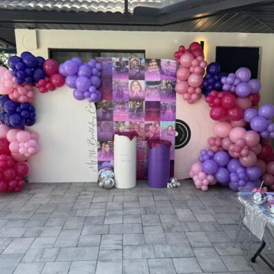 Balloon arch in shades of pink, purple, and red surrounds a photo backdrop and two decorative columns at an outdoor celebration event with a tiled floor—perfect for any Central Florida party shop setup.