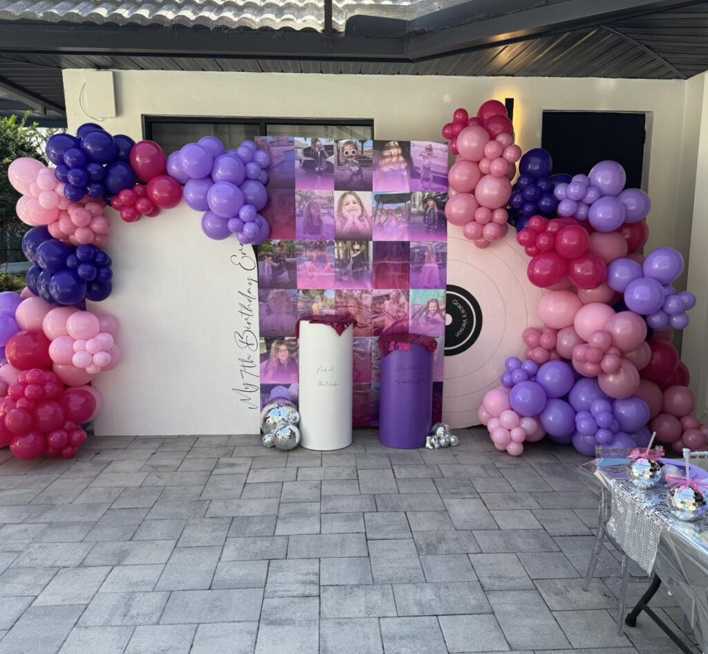 Balloon arch in shades of pink, purple, and red surrounds a photo backdrop and two decorative columns at an outdoor celebration event with a tiled floor—perfect for any Central Florida party shop setup.